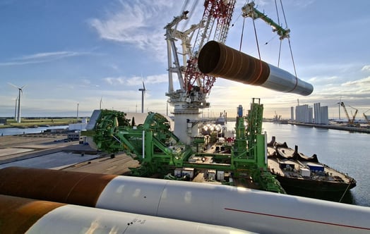 A large crane lifting a monopile foundation at a port, with installation equipment and wind turbines visible nearby. A large crane lifting a monopile foundation at a port, with installation equipment and wind turbines visible nearby.
