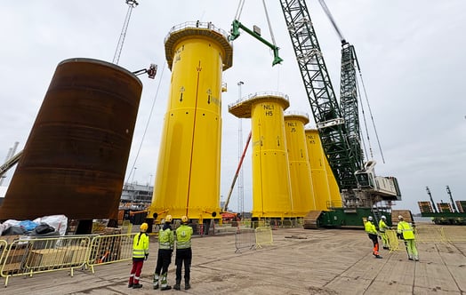 Large offshore wind transition pieces standing in a port area, with cranes and workers nearby.